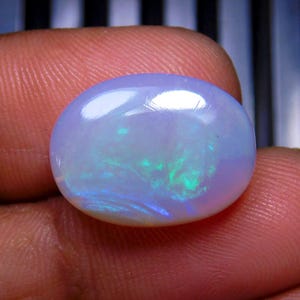 May include: An oval-shaped cabochon opal gemstone featuring a translucent, milky white base with vibrant flashes of iridescent green and blue light play across its smooth, polished surface, showcasing a distinct play-of-color effect throughout.