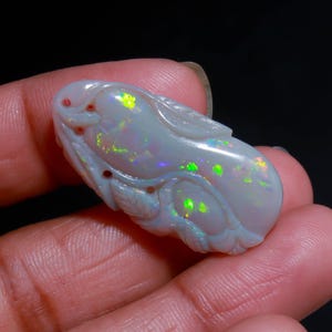 May include: A carved white opal gemstone featuring a smooth, elongated, organic shape with intricate relief details. The surface displays vibrant flashes of green, yellow, and orange play-of-color throughout the polished, translucent stone.