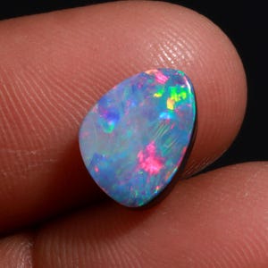 May include: A vibrant, polished opal gemstone with a mix of colors, including blue, pink, and green. The stone has a smooth, rounded shape and a dark base. The opal is held between two fingers, showcasing its iridescent play-of-color.