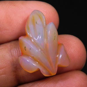 May include: A carved gemstone in the shape of a leaf. The gemstone has a translucent appearance with a mix of colors, including orange, yellow, and hints of purple. The leaf design has detailed edges and a polished finish.