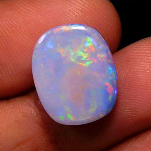 May include: A polished, oval-shaped opal gemstone. The stone has a milky white base color with iridescent flashes of green, blue, and red. The opal's surface is smooth and reflects light, showcasing its play-of-color.
