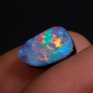 May include: A polished, oval-shaped opal gemstone. The stone displays a vibrant play-of-color, with flashes of blue, green, yellow, and red hues. The opal is set against a dark background, highlighting its iridescent qualities.