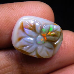 May include: A carved opal gemstone with a flower design. The opal has a white base with brown and orange accents. The carved flower has iridescent green and orange flashes.