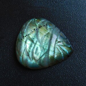 May include: A carved labradorite gemstone with an iridescent sheen. The stone displays detailed, etched patterns in shades of green, blue, and touches of yellow. The gemstone is set against a dark background, accentuating its shimmering qualities.