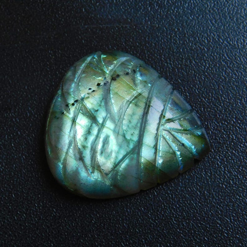 May include: A carved labradorite gemstone with an iridescent sheen. The stone displays detailed, etched patterns in shades of green, blue, and touches of yellow. The gemstone is set against a dark background, accentuating its shimmering qualities.