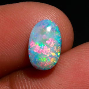 May include: An oval-shaped opal gemstone with a vibrant display of colors. The opal features a base of light blue and green, with flashes of pink, yellow, and orange throughout. The gemstone is held between two fingers.