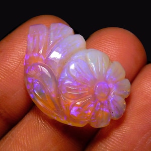 May include: A carved opal gemstone featuring a floral motif with intricate petal detailing. The stone displays a translucent white base color with vibrant flashes of iridescent blue, purple, and orange light throughout its polished surface.