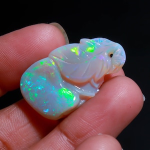 May include: A carved opal gemstone featuring a smooth, rounded base and a sculpted upper section with leaf-like patterns. The stone displays a milky white body color with vibrant, iridescent flashes of neon green and bright blue light.
