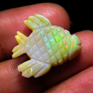 May include: A carved opal gemstone shaped like a fish with a grid-like scale pattern. The stone displays a translucent pale yellow base color with vibrant flashes of green and orange iridescence across its surface, highlighting the carved details.