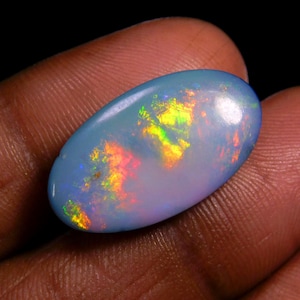 Puede incluir: Gema de ópalo ovalada con un color base azul claro. El ópalo muestra un vibrante fuego interno con destellos amarillos, naranjas, rojos y verdes. La gema se sostiene en una mano sobre un fondo oscuro.