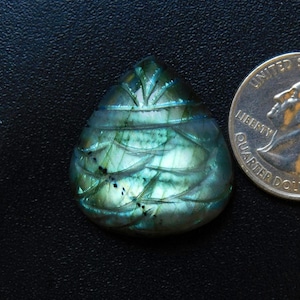May include: A teardrop-shaped, carved labradorite gemstone with iridescent blue, green, and teal hues. The stone features intricate carved details and is next to a U.S. quarter for size comparison. The gemstone is set against a dark background.