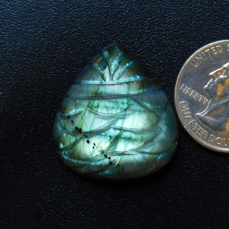 May include: A teardrop-shaped, carved labradorite gemstone with iridescent blue, green, and teal hues. The stone features intricate carved details and is next to a U.S. quarter for size comparison. The gemstone is set against a dark background.