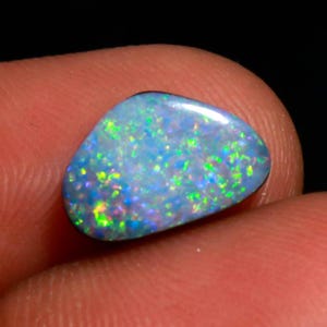 May include: A polished, oval-shaped opal gemstone. The opal displays a vibrant play-of-color, with flashes of green, blue, and yellow hues set against a light blue base. The gemstone is held between fingers, showcasing its natural beauty.