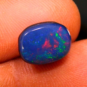 Puede incluir: Una piedra preciosa de ópalo negro pulido, de forma ovalada. El ópalo muestra un color base azul oscuro con vibrantes destellos de rojo, verde y azul en su superficie. La piedra preciosa se sostiene entre dos dedos.