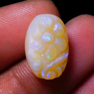 May include: Oval gemstone with a carved floral pattern. The stone has a creamy yellow base colour with iridescent blue and green flashes. The carving is detailed, showcasing the flower's petals.