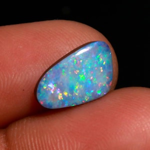May include: A polished, oval-shaped opal gemstone. The opal displays a vibrant play-of-color, with flashes of blue, green, and red hues across a milky white base. The gemstone is held between two fingers against a dark background.