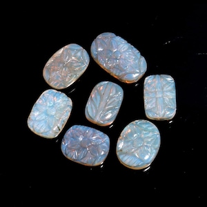 May include: A collection of carved opalescent gemstones with floral and leaf designs. The gemstones are in various shapes, including oval, rectangular, and square, and are arranged on a black surface. The carvings are detailed and intricate, with a soft, ethereal glow.