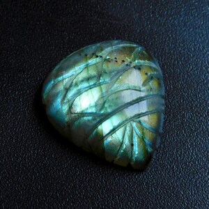 May include: A polished labradorite gemstone with carved leaf-like patterns. The stone displays iridescent colours of blue, green, and gold. The gemstone is set against a dark background.
