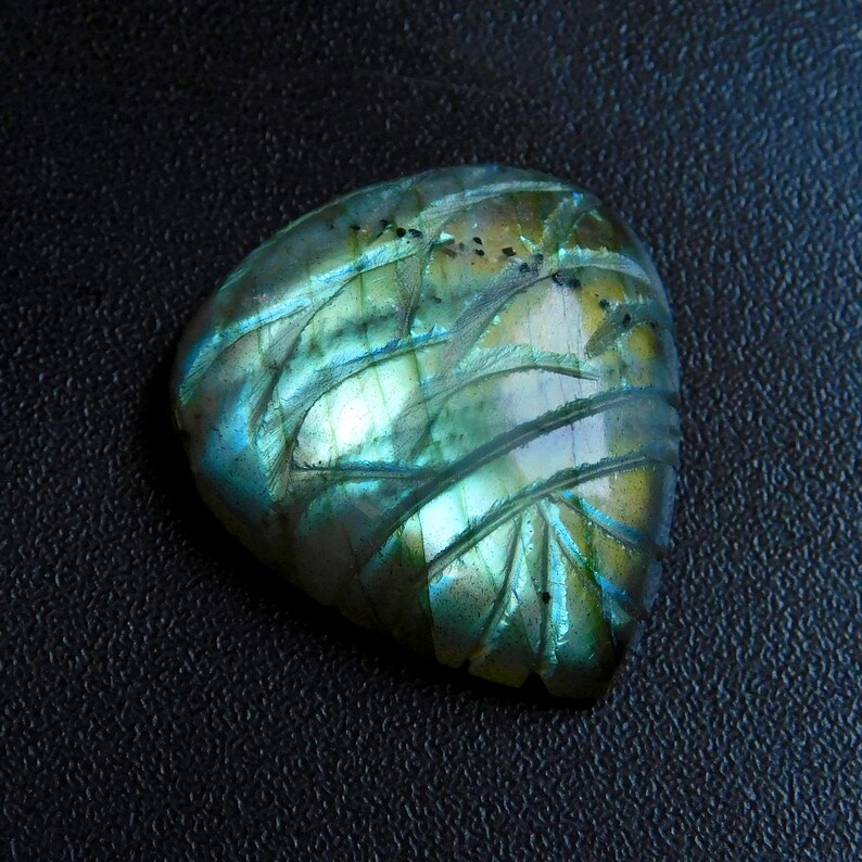 May include: A polished labradorite gemstone with carved leaf-like patterns. The stone displays iridescent colours of blue, green, and gold. The gemstone is set against a dark background.