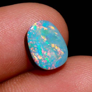 May include: A polished, oval-shaped opal gemstone. The opal displays a vibrant play-of-color, with flashes of blue, pink, and yellow hues. The gemstone is held between two fingers against a dark background, highlighting its iridescent qualities.