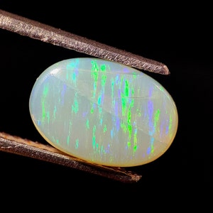 Könnte beinhalten: Ein weißer ovaler Opal-Edelstein mit grünen und blauen irisierenden Blitzen. Der Opal wird in einer Pinzette vor einem schwarzen Hintergrund gehalten.
