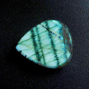 May include: A polished, teardrop-shaped labradorite gemstone. The stone has a vibrant play of colour, with iridescent teal and blue hues. Darker, linear patterns cross the surface, creating a grid-like effect. The gemstone is set against a dark background.