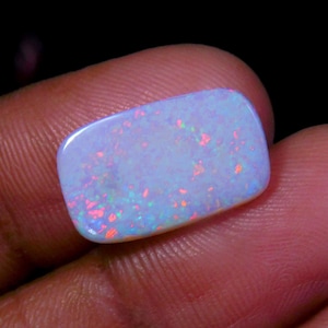 Puede incluir: Una piedra preciosa de ópalo rectangular pulida. El ópalo tiene un color base azul pálido con destellos rojos, naranjas y verdes. La piedra se sostiene entre los dedos sobre un fondo oscuro.