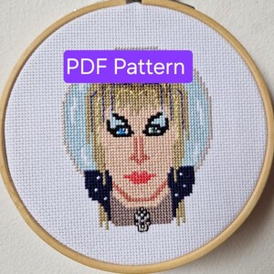 May include: Cross-stitch portrait of a person with blonde hair, blue eyes, and red lips, set in a wooden embroidery hoop. The design includes blue accents and a black collar. The text "PDF Pattern" is displayed in a purple rectangle.