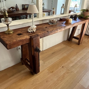 Può includere: Un lungo banco da lavoro rettangolare in legno caratterizzato da una spessa superficie marrone usurata con venature e texture visibili. Il tavolo include una maniglia a morsa in metallo montata lateralmente e robuste gambe inclinate.