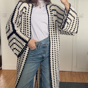 Peut inclure: Longue veste en crochet ouverte sur le devant, ornée d'un motif géométrique crème et bleu marine. La veste est portée sur un t-shirt blanc et un jean bleu. Les manches sont larges et la veste descend jusqu'aux chevilles.