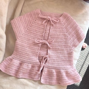 以下が含まれることがあります： フリルのある裾と首元にリボンが付いたピンクのクロシェトップ