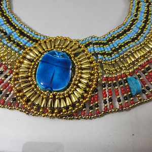May include: A gold and blue beaded necklace with a large blue scarab beetle pendant. The necklace is made of multiple rows of beads and has a fringe of red, black, and gold beads.