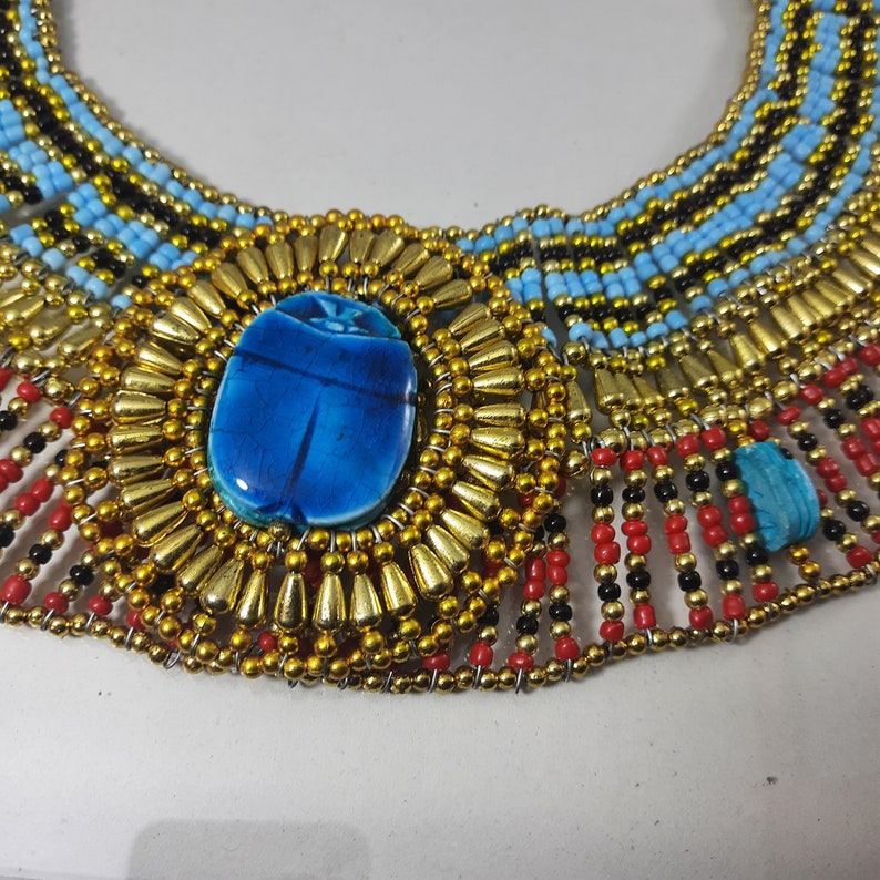 May include: A gold and blue beaded necklace with a large blue scarab beetle pendant. The necklace is made of multiple rows of beads and has a fringe of red, black, and gold beads.