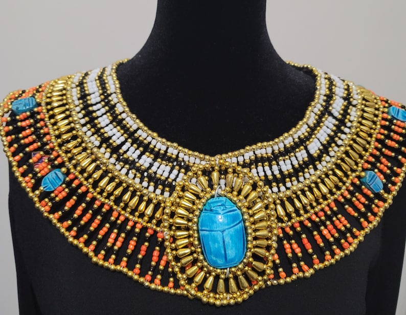 May include: A gold and black beaded necklace with a turquoise scarab beetle pendant. The necklace is layered with different sizes of beads and has a fringe of gold beads.