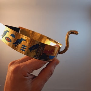 May include: A gold-colored headband with a cobra design and Egyptian hieroglyphs. The headband is decorated with blue, red, and black enamel.