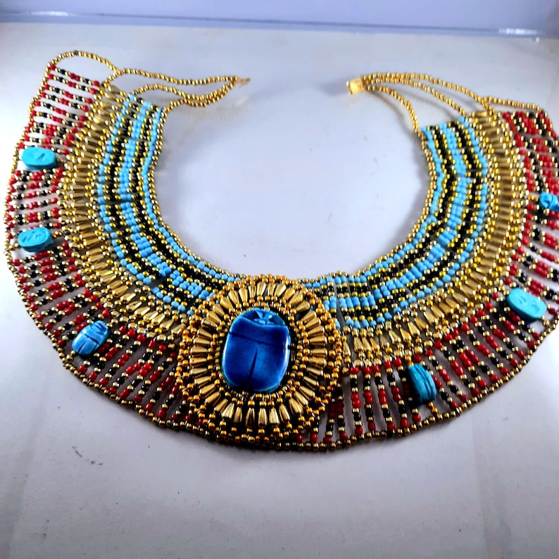 May include: A gold and blue beaded necklace with a large turquoise scarab beetle pendant. The necklace is made of small beads in red, black, and turquoise colors.