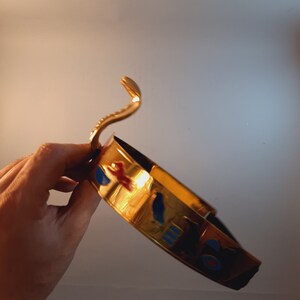 May include: A gold-colored crown with blue, red, and white accents. The crown features a snake design on the top.