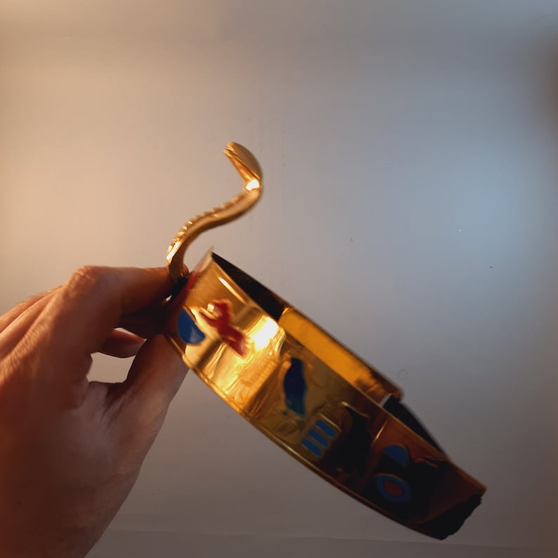 May include: A gold-colored crown with blue, red, and white accents. The crown features a snake design on the top.
