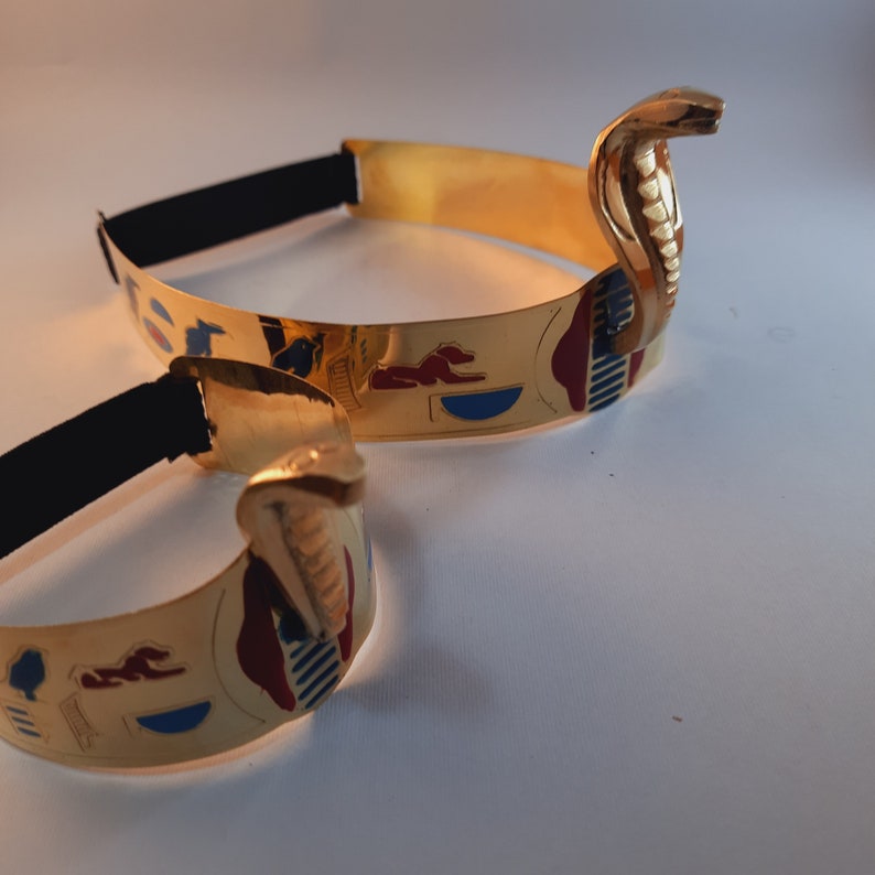 May include: Two gold-colored headbands with an Egyptian-inspired design. Each headband features a cobra head with blue, red, and gold accents. The headbands are made of metal and have an elastic band for a comfortable fit.