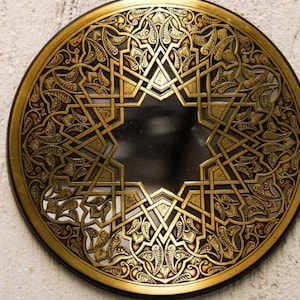 May include: A round, gold-colored metal wall decor with an intricate, geometric design. The design features a central star shape surrounded by a repeating pattern of leaves and swirls.