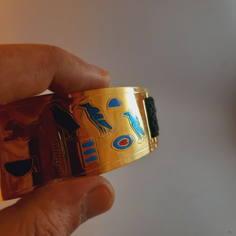 May include: Gold-toned bracelet with a black band and an intricate design featuring blue and red enamel accents. The design includes hieroglyphics and stylized birds.