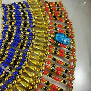 May include: A beaded necklace with gold, red, blue, and black beads. A blue scarab beetle charm hangs from the center of the necklace.