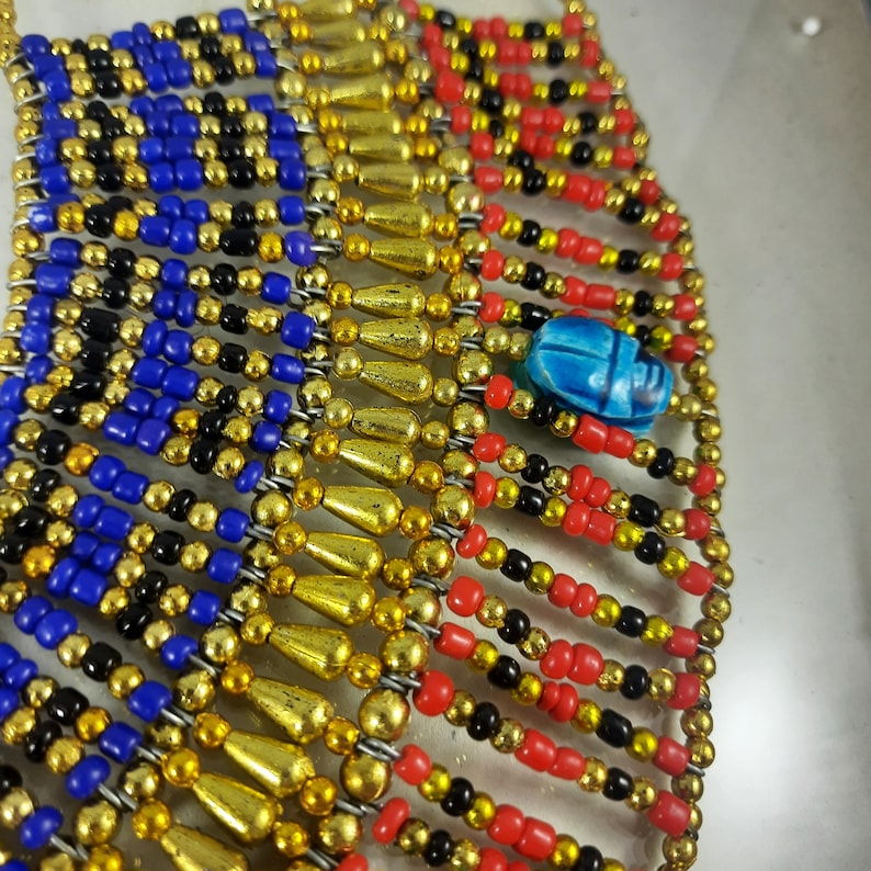 May include: A beaded necklace with gold, red, blue, and black beads. A blue scarab beetle charm hangs from the center of the necklace.