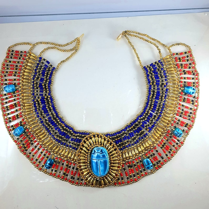 May include: A gold, blue, red, and black beaded necklace with a large blue scarab beetle pendant. The necklace is in the style of an ancient Egyptian collar.