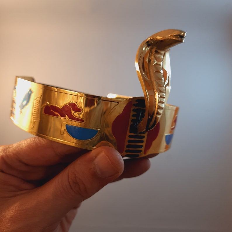May include: Gold-toned bracelet with a raised cobra design. The bracelet is decorated with colorful enamel and hieroglyphic symbols.