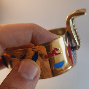 May include: Gold-toned bangle bracelet with an Egyptian-inspired design. The bracelet features colorful enamel details, including a blue circle, red and white stripes, and a red lion. The bracelet has a hinged opening.