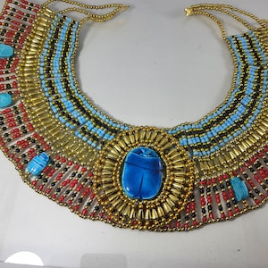 May include: A gold and blue beaded necklace with a large blue scarab beetle pendant. The necklace is made of small beads in red, black, and gold colors.