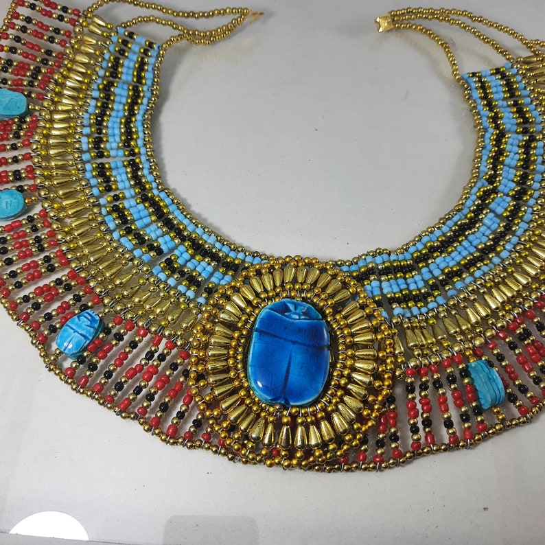 May include: A gold and blue beaded necklace with a large blue scarab beetle pendant. The necklace is made of small beads in red, black, and gold colors.