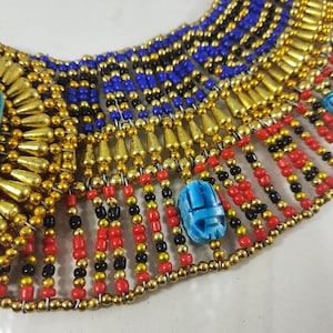 May include: A gold and blue beaded necklace with a scarab beetle charm. The necklace is made of small, round beads in shades of gold, blue, red, and black. The scarab beetle charm is made of blue glass and is set in a gold frame.