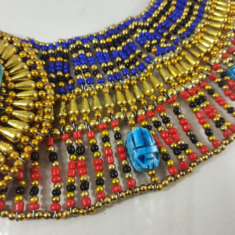 May include: A gold and blue beaded necklace with a scarab beetle charm. The necklace is made of small, round beads in shades of gold, blue, red, and black. The scarab beetle charm is made of blue glass and is set in a gold frame.